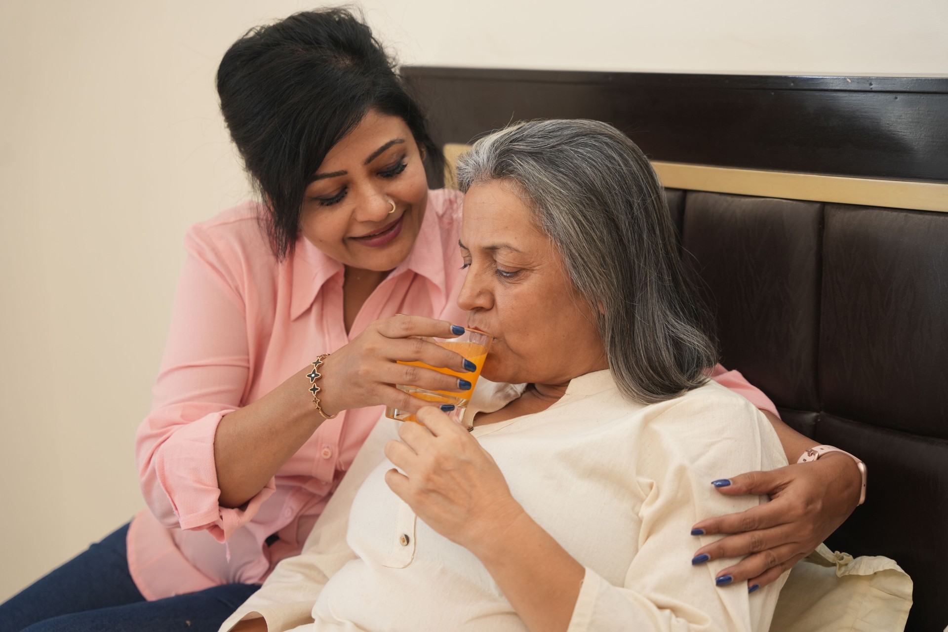 young indian daughter helping her senior mother drink orange juice not well. healthcare and recovery concept.